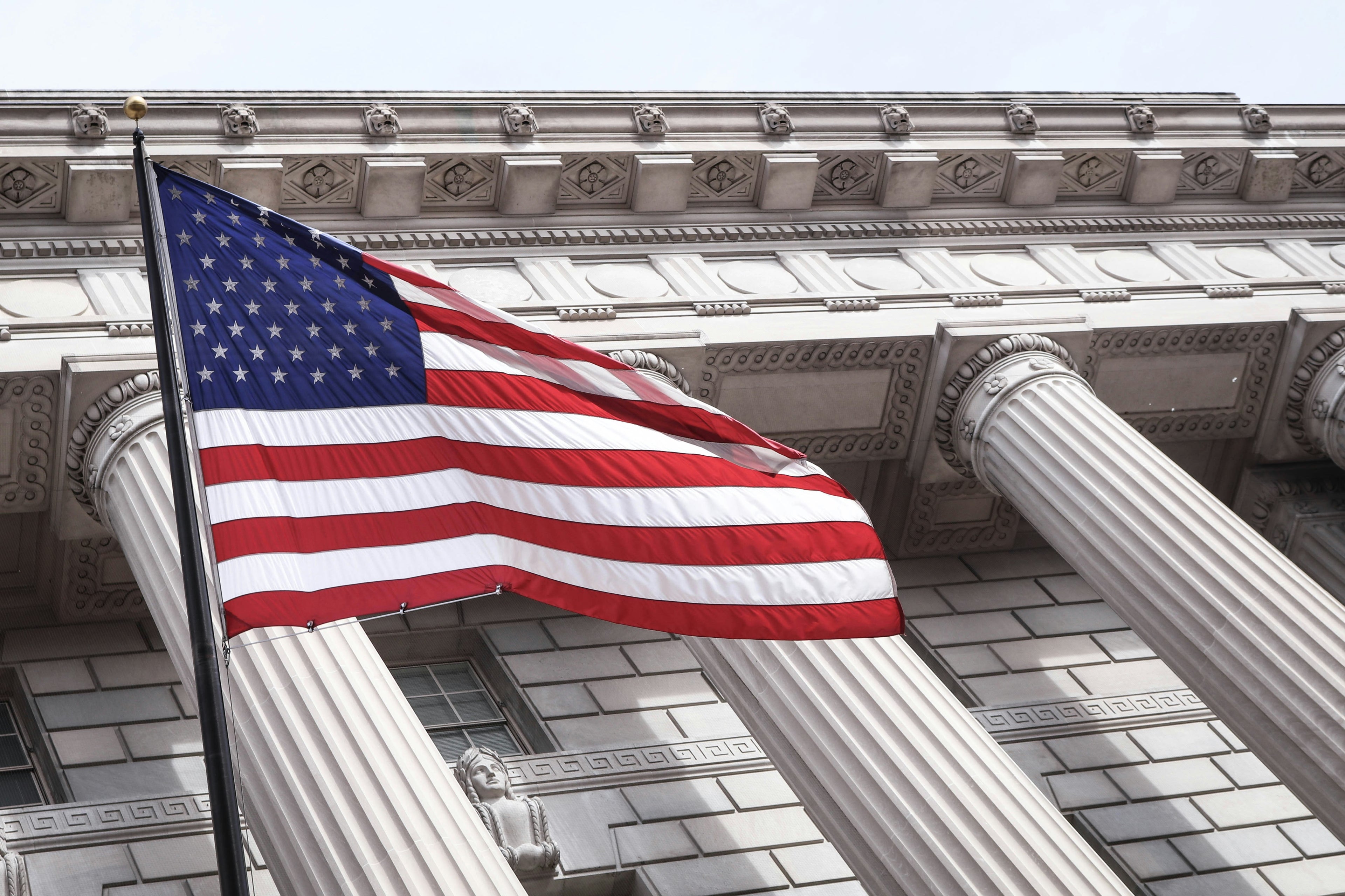 Photo of U.S. flag flying on the left in front of  pillars on the US capitol building. Photo by Brandon Mowhinkel vis Unspplash